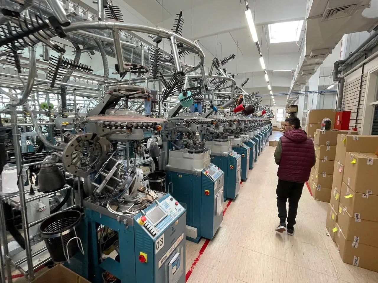 A knitting-hall aisle with a row of teal circular knitting machines; a worker in a maroon jacket walks down the aisle past stacked cardboard shipping boxes.
