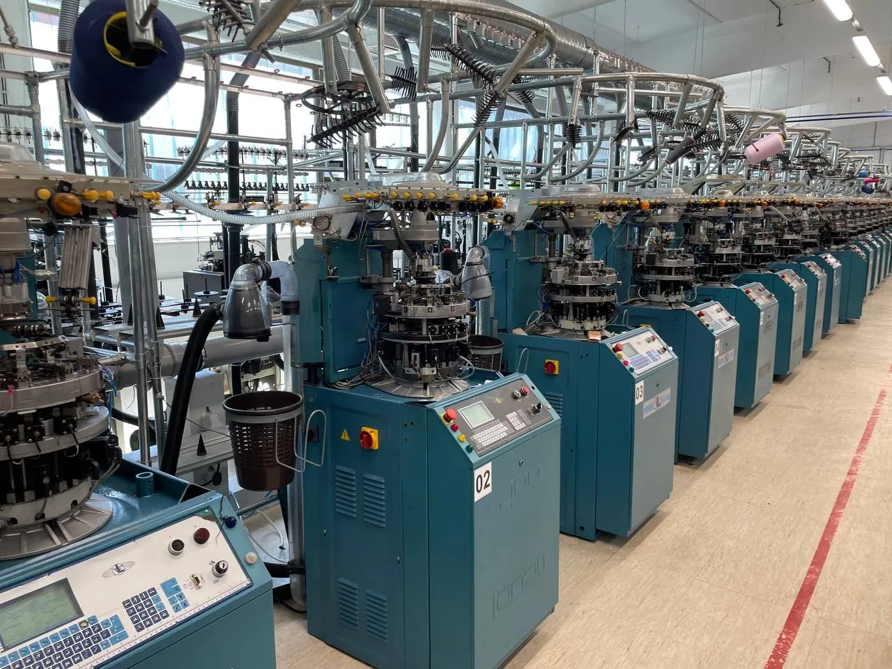 View down a knitting-hall aisle with a long row of teal circular knitting machines and yarn cones and guide arms suspended overhead, with no worker in frame.