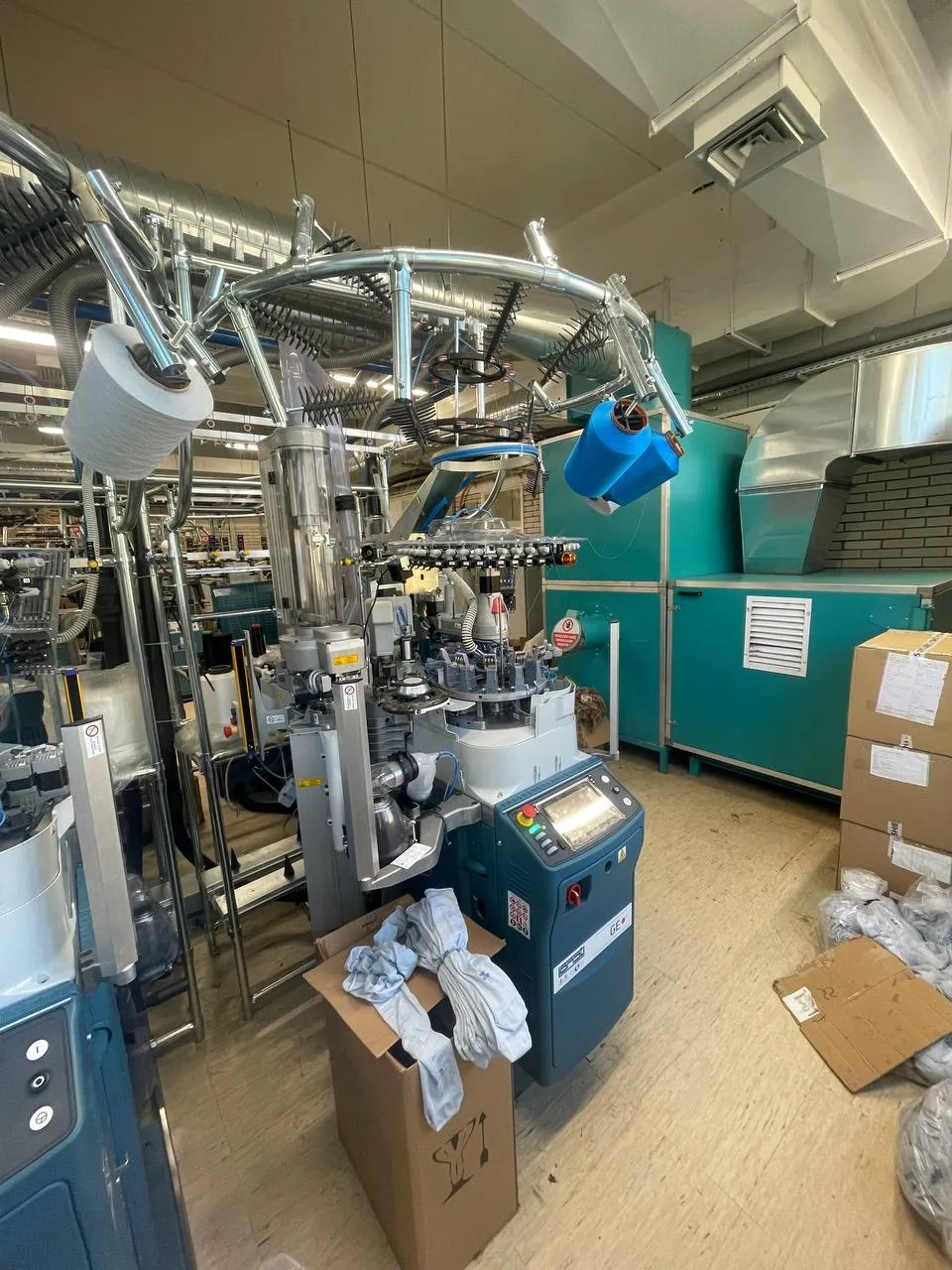 Close-up of a knitting machine with large cones of white and blue yarn above the control panel, and socks draped over a cardboard box in the foreground.