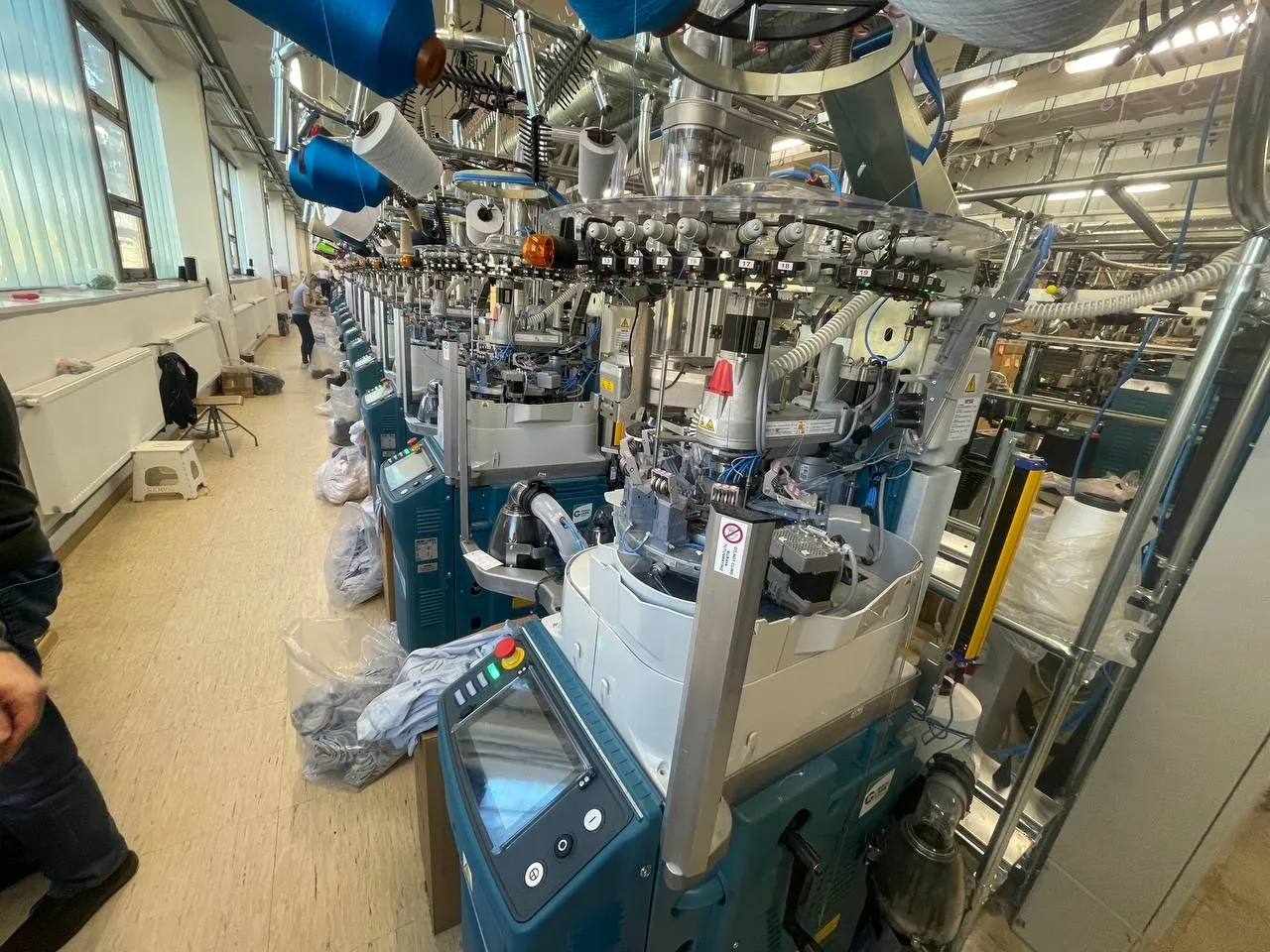 Close-up of a circular knitting machine's needle cylinder and yarn feed, with a long row of machines behind it and a worker visible near the windows.