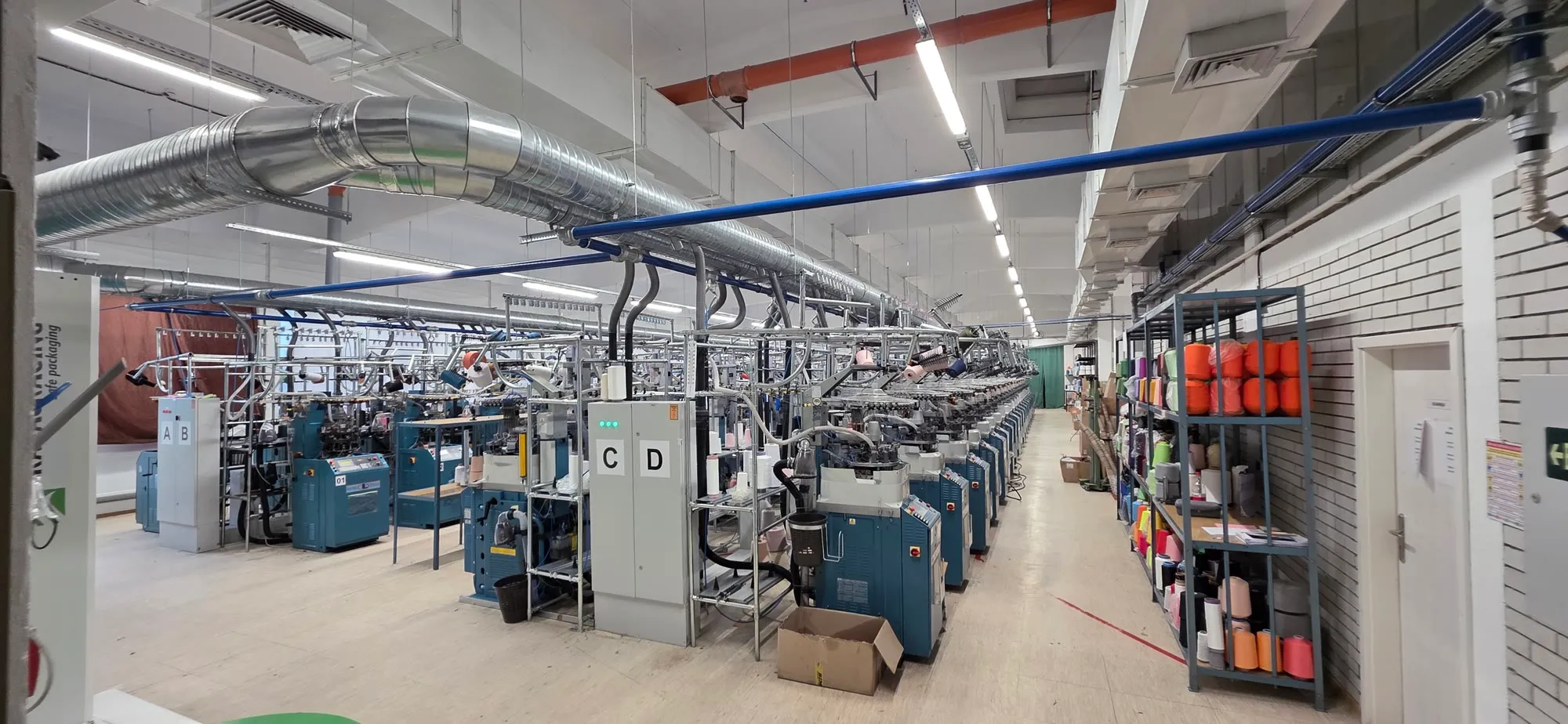 Wide view of a knitting hall with teal knitting machines, overhead ducting, and a shelf of coloured yarn cones against the right-hand wall.