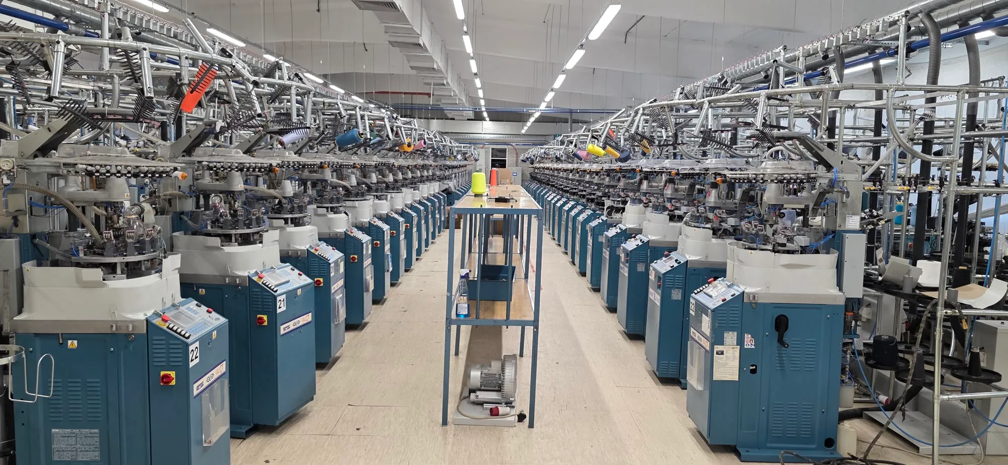 A long central aisle between two rows of teal circular knitting machines, with yarn guides overhead and a work table in the middle.