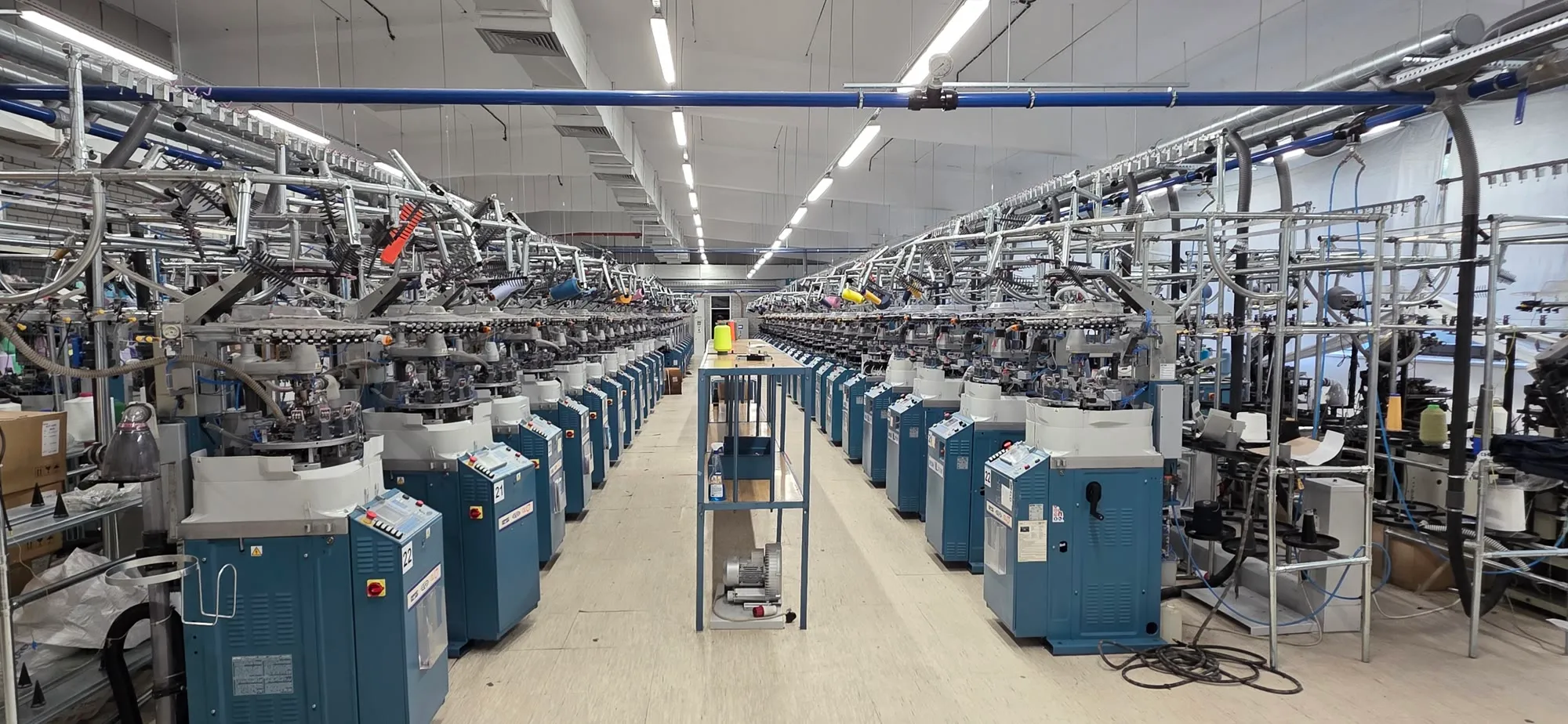 Two rows of teal circular knitting machines stretching into the distance on either side of an aisle, under bright ceiling lights.