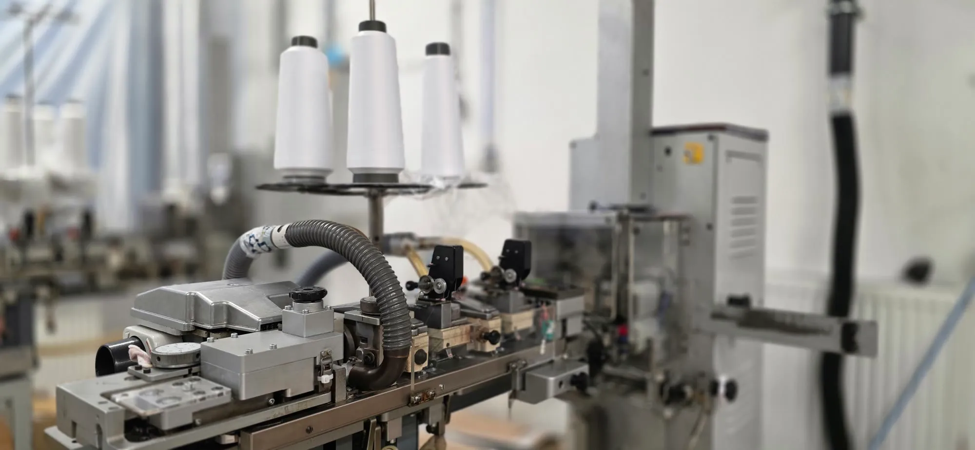 A toe-seaming machine with three white thread cones on top and its conveyor and suction hose in the foreground.